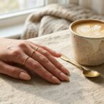 Elegant nude nails manicure flat lay with cozy linen fabric, ceramic latte cup, and luxury gold rings