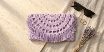 Lavender recycled cotton crochet clutch bag pattern with semi-circle lacy flap and fringe on travertine stone