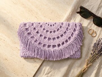 Lavender recycled cotton crochet clutch bag pattern with semi-circle lacy flap and fringe on travertine stone