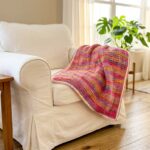 Puff stitch crochet blanket in variegated pink coral and gold yarn draped over a white armchair