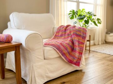 Puff stitch crochet blanket in variegated pink coral and gold yarn draped over a white armchair