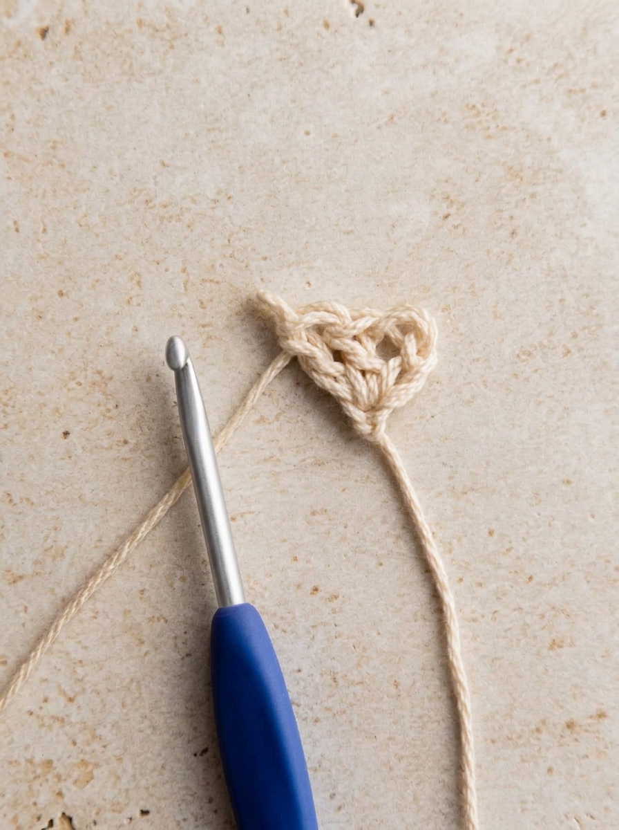 Row 1 of crochet bandana pattern showing first 3-dc granny cluster in cream cotton yarn with blue hook